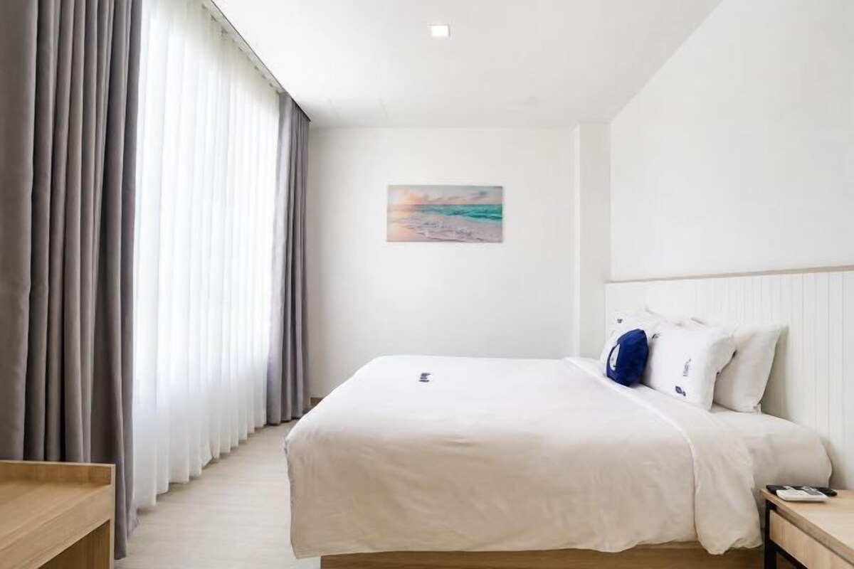 Een hotelkamer met een bed, nachtkastje, schilderij aan de muur en een raam met gordijnen.