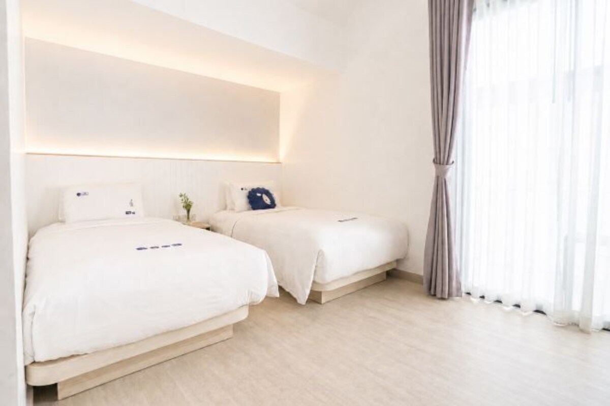Habitación de hotel con dos camas, una mesita de noche con lámpara y un ventanal con cortinas.