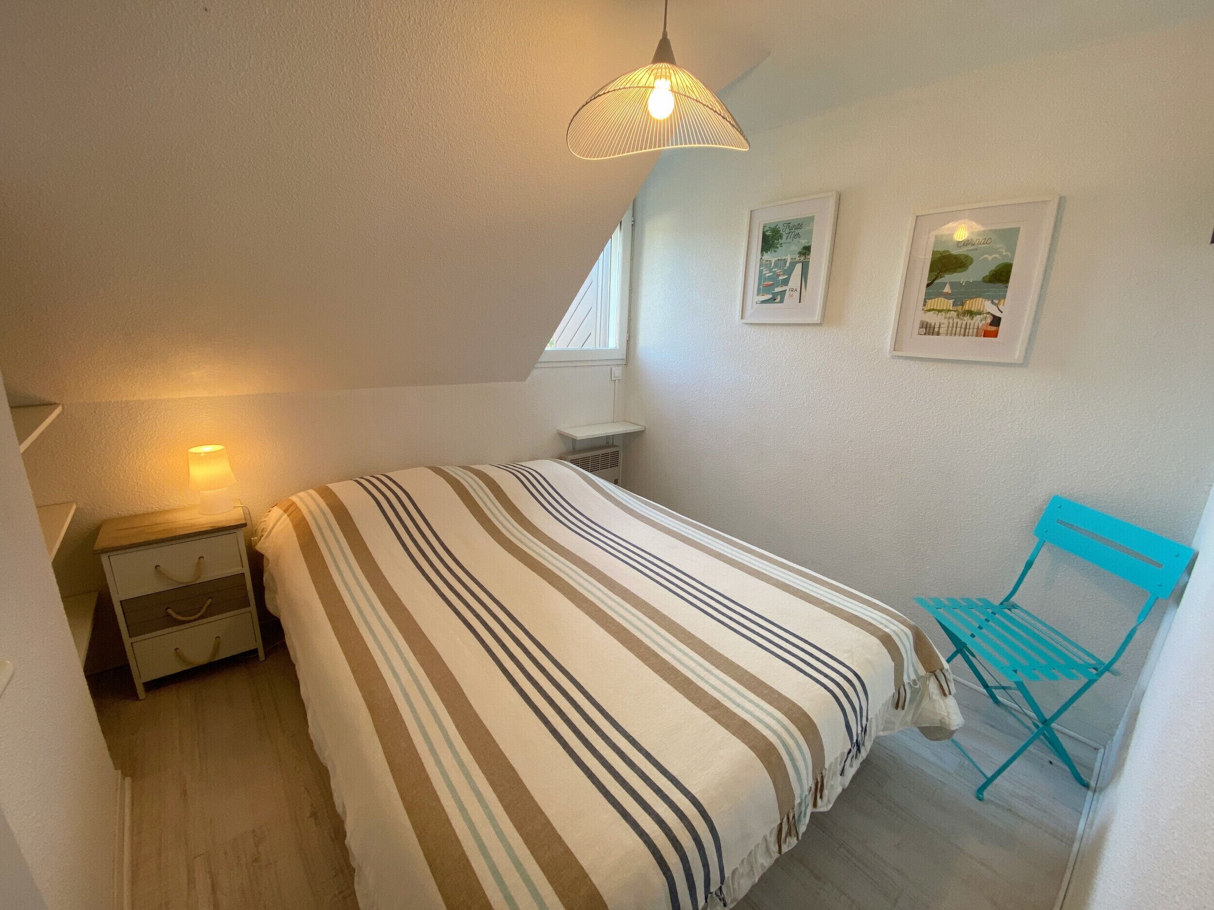 Een slaapkamer met een bed, nachtkastje, lamp, raam en stoel.