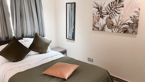 A bedroom with a bed, pillows, a nightstand, a wall-mounted mirror, and a tropical-themed painting. - Beaufort Ridge (Accra)