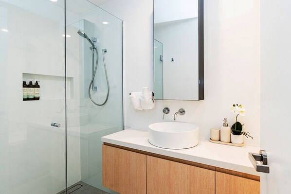 A modern bathroom with a glass shower enclosure, a white sink, and a wooden vanity. - Beachfront Escape at Beach Life with Ocean Views (Alexandra Headland)