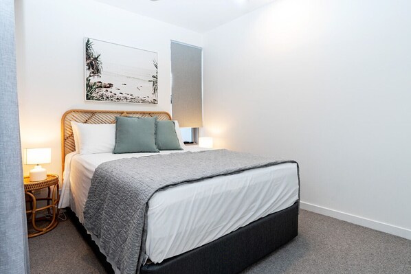 Quarto bem arrumado com cama, duas luminárias de cabeceira, uma obra de arte na parede e uma mesinha de apoio.