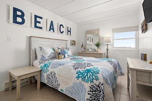 Une chambre à coucher comprenant un lit, une table de chevet, une commode, un miroir et un mur sur lequel est inscrit le mot « PLAGE ».