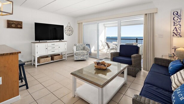 A living room with a TV, white furniture, a glass coffee table, wicker chairs, and a balcony with a view. - Oceanfront Three Bedroom Condo on the Boardwalk (Ocean City)