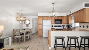 A modern kitchen with wooden cabinets, a white refrigerator, a microwave, and a granite countertop. There are three bar stools and a dining table with chairs. - Oceanfront Three Bedroom Condo on the Boardwalk (Ocean City)