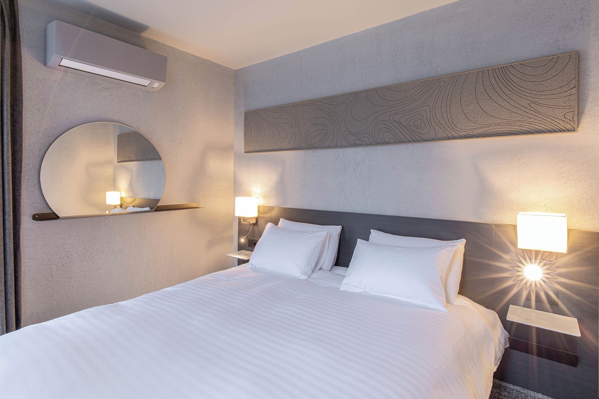 A hotel room with a large bed, two bedside tables with lamps, a wall-mounted air conditioner, a round mirror, and a decorative wall panel.
