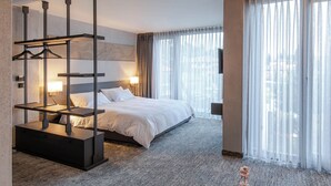A hotel room with a large bed, a bedside table with a lamp, a wardrobe with a mirror, and a view of the city through sheer curtains. - Therma Numera Longevity SPA (Burgas)
