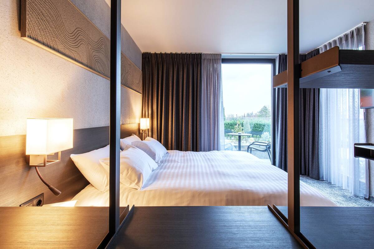 A hotel room with a large bed, two bedside lamps, a view of the outdoors through a window, and a wooden headboard.