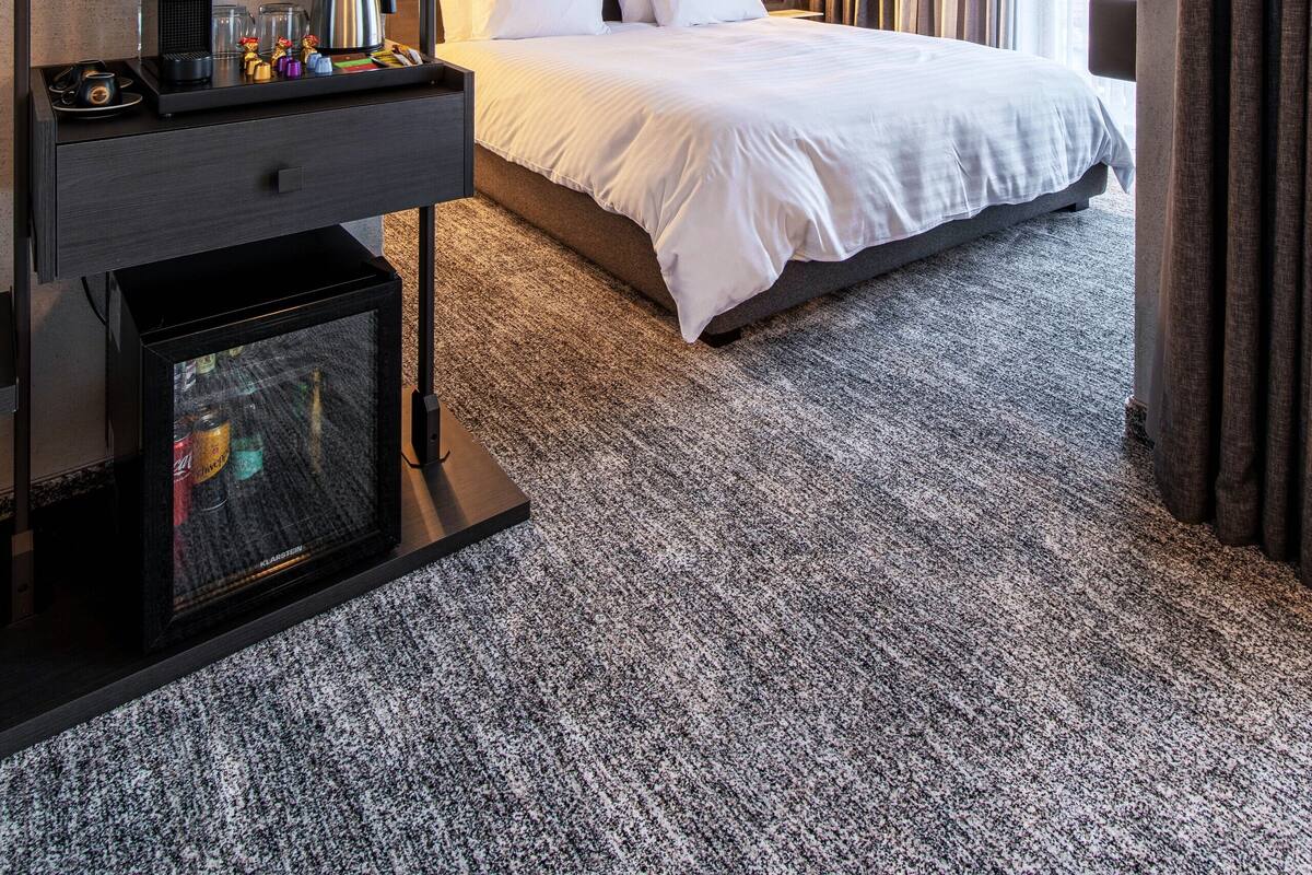 A hotel room with a bed, bedside table, lamp, and a view of the outdoors.