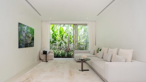 Un salon moderne avec un canapé blanc, une table basse et une grande fenêtre donnant sur des plantes tropicales.
