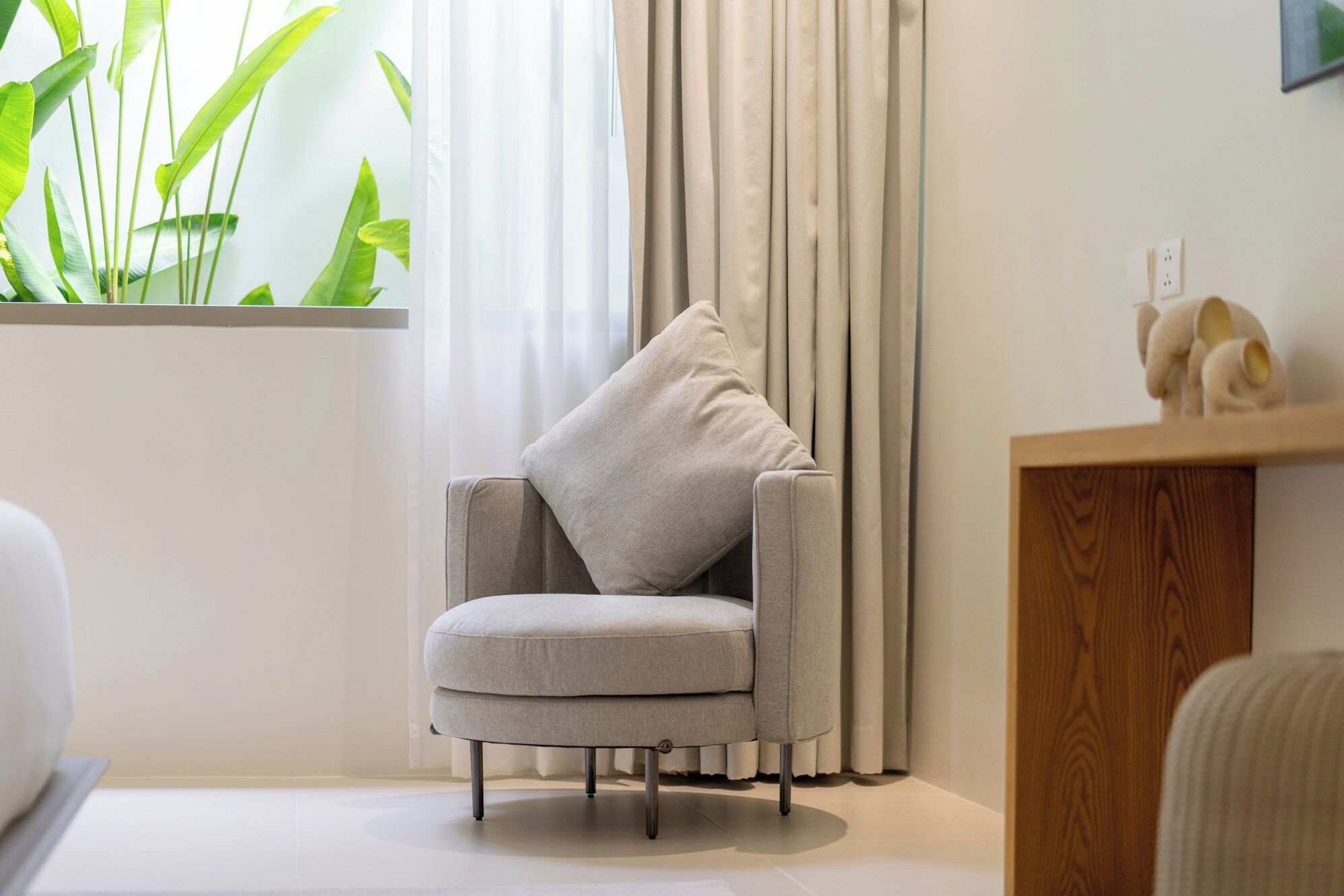 Un sillón moderno con un cojín, situado cerca de una ventana con cortinas translúcidas y una planta verde.