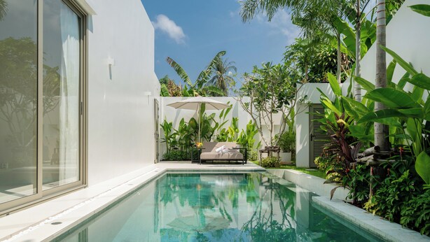 Une piscine dotée d’un mur en verre, entourée d’une végétation luxuriante et bordée d’un bâtiment blanc.