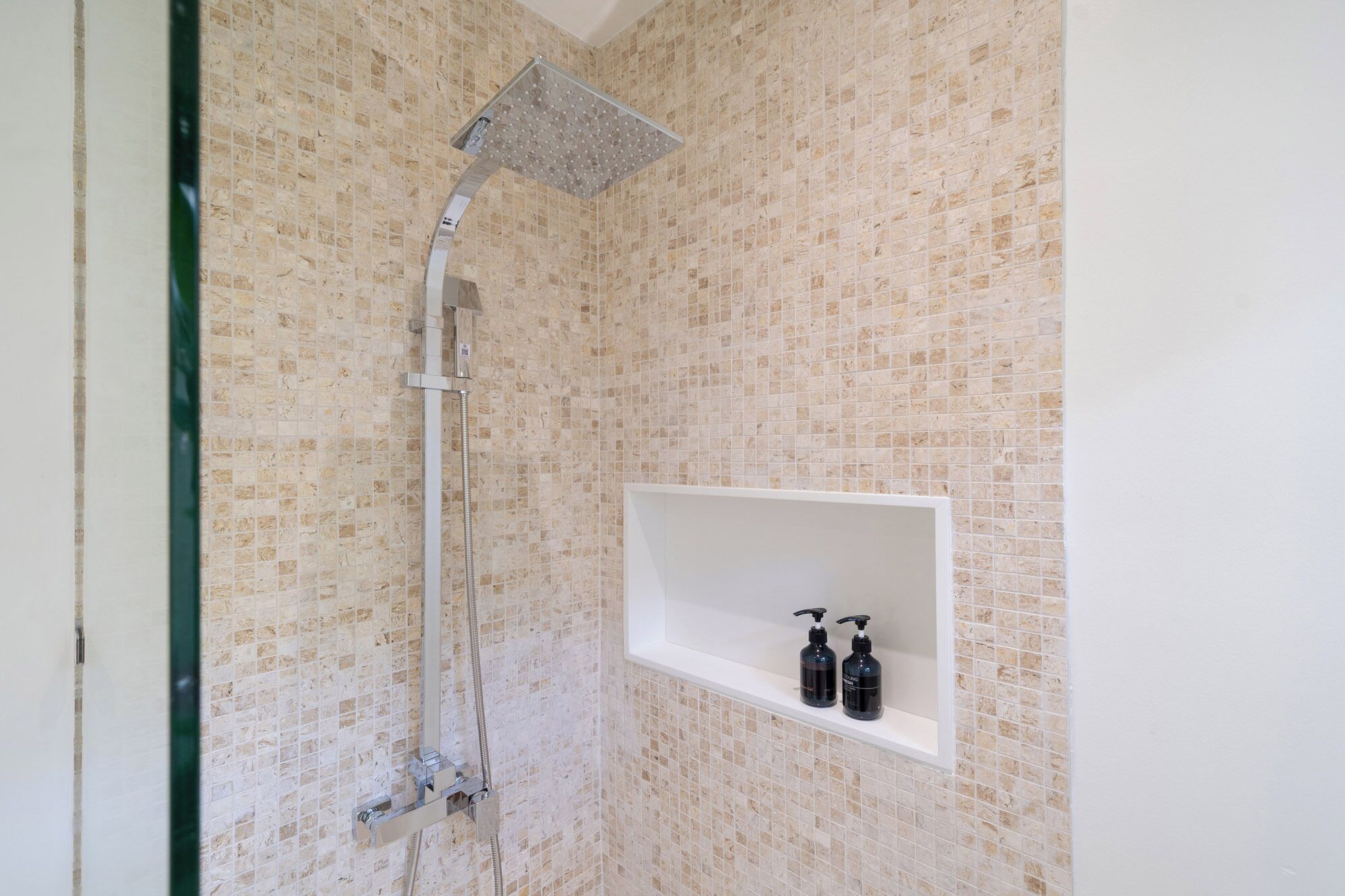 A shower with a square head, a wall-mounted shelf with two bottles, and a tiled wall.