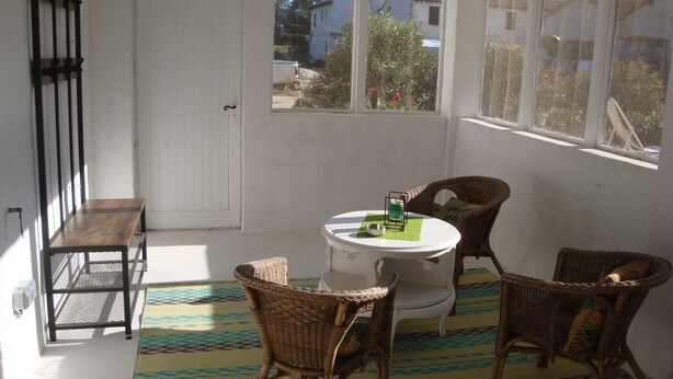 Una habitación soleada con una puerta blanca, sillas de mimbre, una mesita con una vela verde y una alfombra estampada.