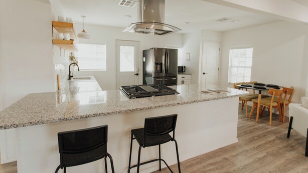 A modern kitchen with a central island, black bar stools, a dining area with wooden chairs, and a stainless steel refrigerator.