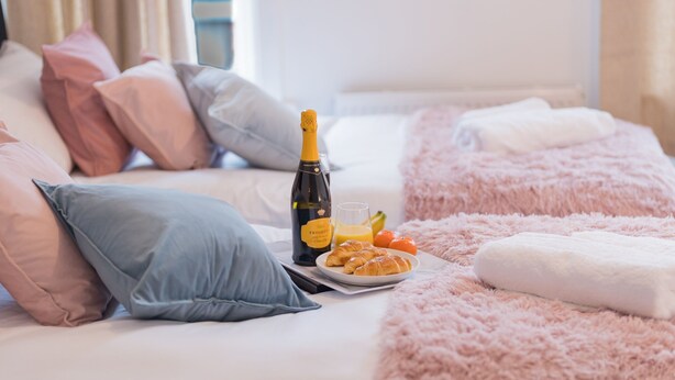A neatly made bed with a fluffy pink blanket, a bottle of champagne, and a plate of croissants and orange juice.