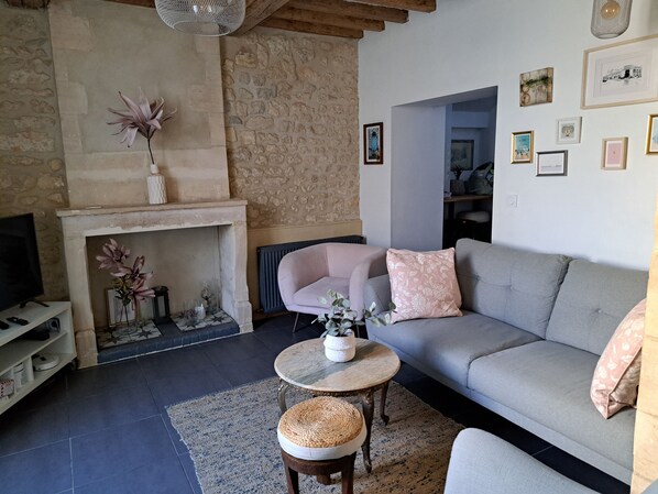 A living room with a stone wall, a fireplace, a grey sofa, a round coffee table, and a television on a stand. - La Petite Bajocasse, a charming townhouse in the center of Bayeux (Bayeux)