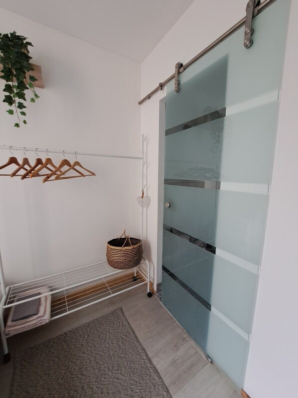 A room with a white wall, a wooden hanger rack, a woven basket, a metal bench, and a sliding glass door. - La Petite Bajocasse, charming town house in downtown Bayeux (Bayeux)