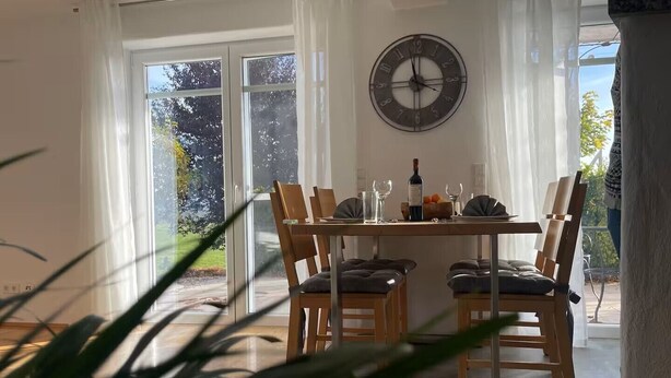 A dining area with a table set for two, a clock on the wall, and a view of the outdoors through large windows. - Holiday apartment - nature, peace and relaxation (Wangen im Allgäu)