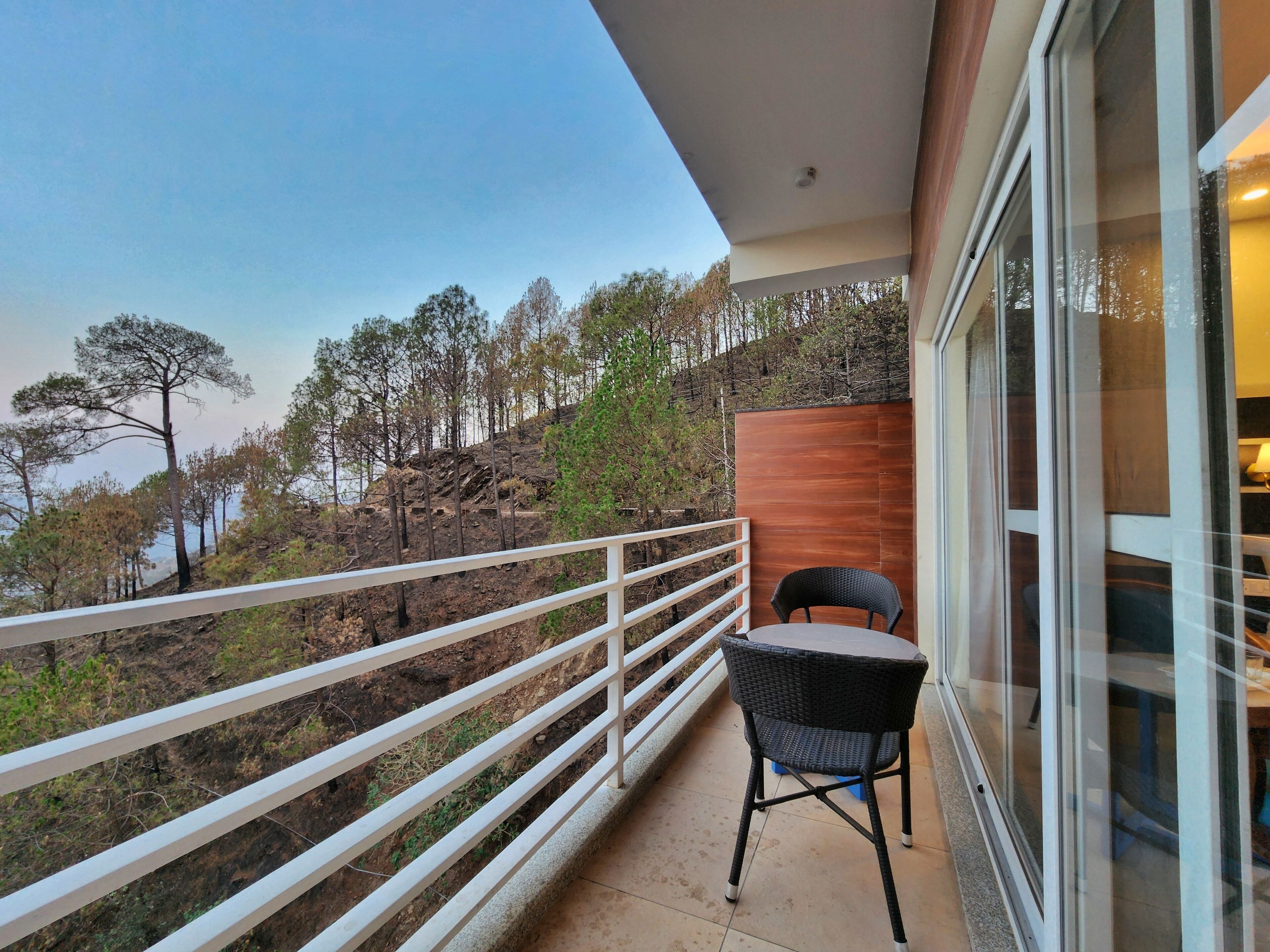 Een balkon met een witte balustrade, een zwarte rieten stoel en uitzicht op bomen en een heuvel.