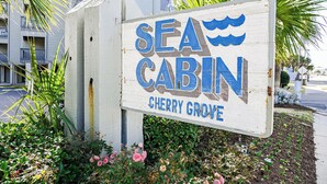 Un letrero de madera que dice 'Sea Cabin' con temática náutica, situado en una zona residencial con un edificio y vegetación de fondo.