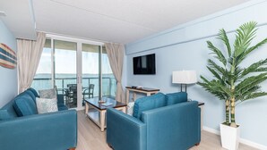 A modern living room with a balcony, a flat-screen TV, and a potted plant. - Sunset Chaser’s Dream | Bayfront Condo Near the Boardwalk (Ocean City)