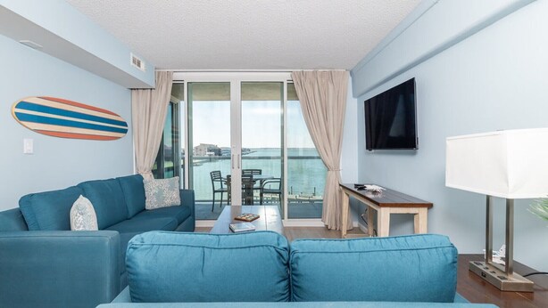 A modern living room with a blue sofa, a small table, a TV, and a view of the sea through large windows. - Sunset Chaser’s Dream | Bayfront Condo Near the Boardwalk (Ocean City)