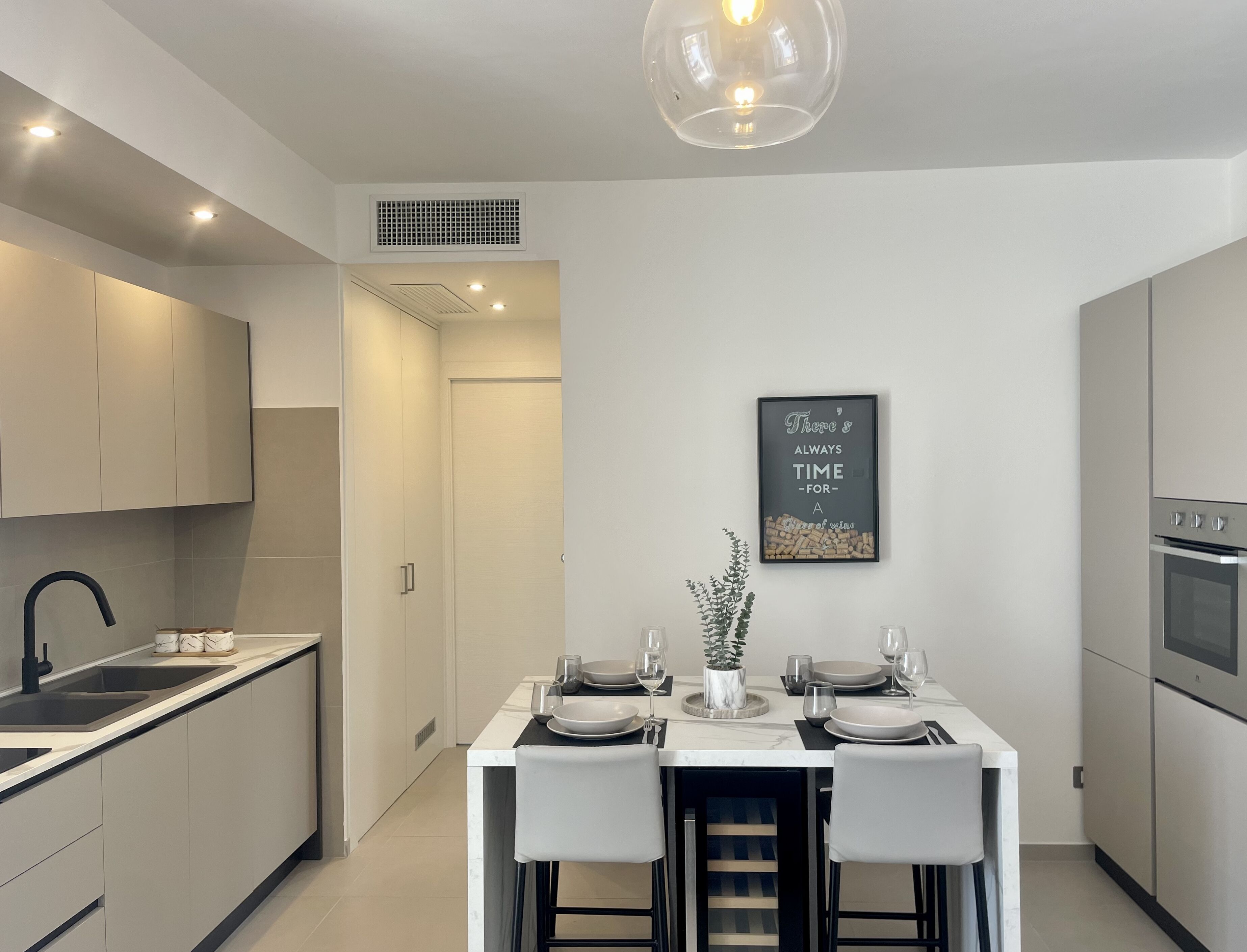 A modern kitchen with a dining area, featuring a central island with chairs, a built-in oven, and a wall-mounted light fixture.