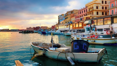 A harbor with boats docked, buildings along the waterfront, and a sunset in the background.