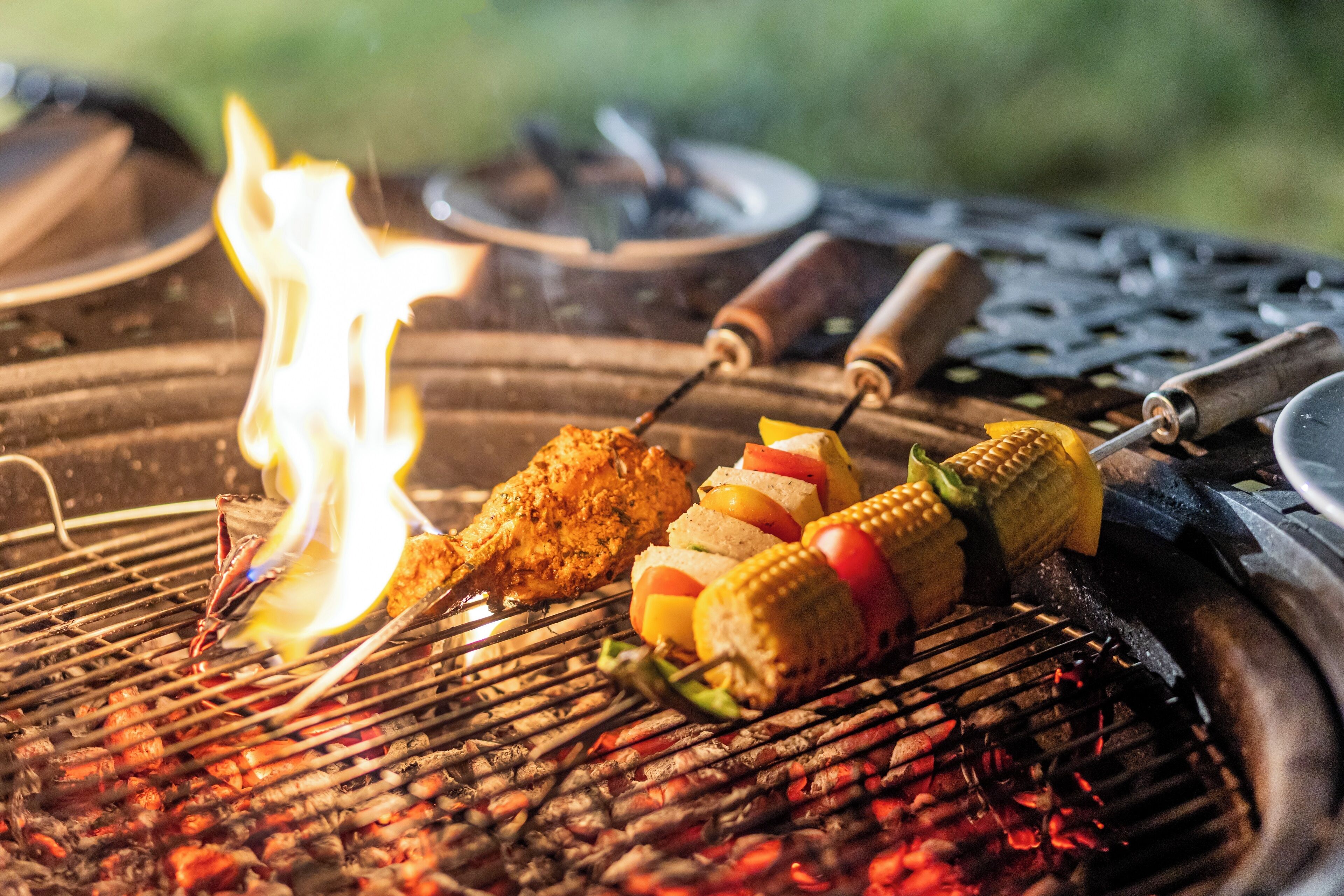 En grill med spyd fyldt med mad, herunder majs og grøntsager, samt en brændende ild.