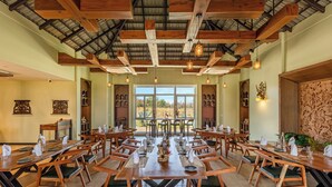 A dining area with wooden tables and chairs, large windows, and a high ceiling with exposed beams. - Sterling Tipeshwar (Kelapur)