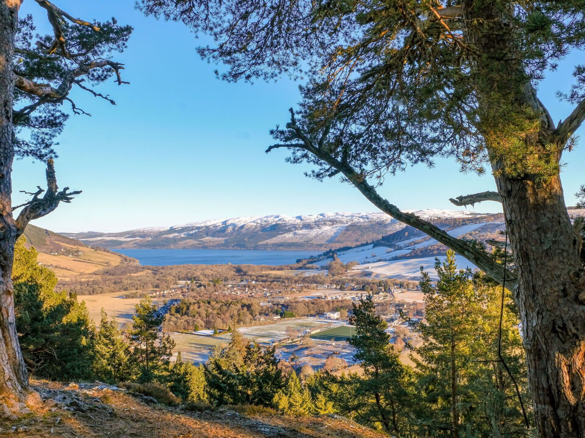 A panoramic view of a mountainous landscape with a lake, surrounded by trees and a small settlement.