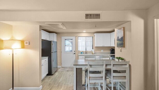 A modern kitchen with white cabinets, a black refrigerator, a dining area with white chairs, and a floor lamp. - Elegant Green Valley Villa 55+ (Green Valley)