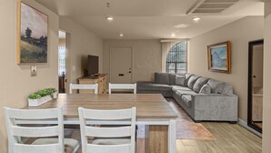 A modern living room with a wooden dining table, white chairs, a grey sectional sofa, and a large painting on the wall. - Elegant Green Valley Villa 55+ (Green Valley)