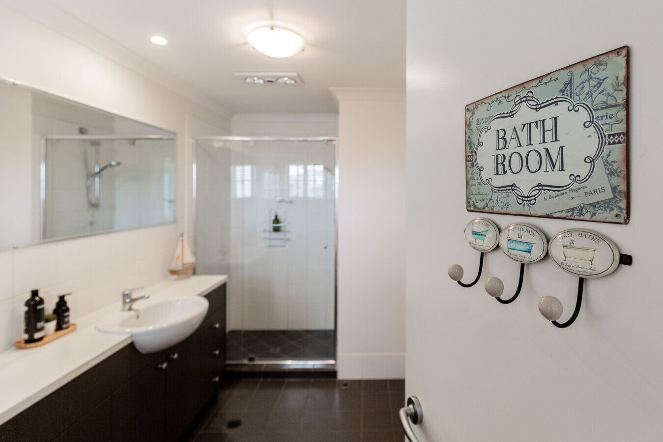 Ein modernes Badezimmer mit einem großen Spiegel, zwei Waschbecken, einer Dusche und einem an der Wand befestigten Schild mit der Aufschrift 'BATH ROOM'.