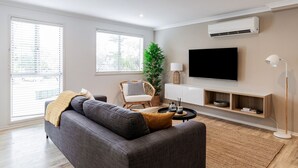 A modern living room with a grey sofa, a small side table, a floor lamp, a TV, and a large window with blinds. - Marcoola Sands Retreat Steps from Marcoola Beach (Marcoola)
