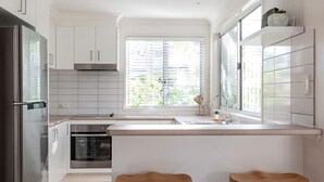 A modern kitchen with white cabinets, a stainless steel refrigerator, a built-in oven, a window with blinds, and two wooden stools. - Marcoola Sands Retreat Steps from Marcoola Beach (Marcoola)
