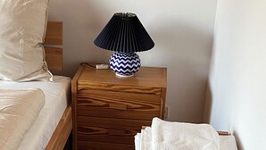 A neatly arranged bedroom with a bed, a wooden nightstand with a lamp, a small table with folded towels, and a dark rug on the floor.