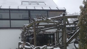 A snow-covered outdoor area with a wooden pergola structure, a stone wall, and a building with a glass facade in the background.