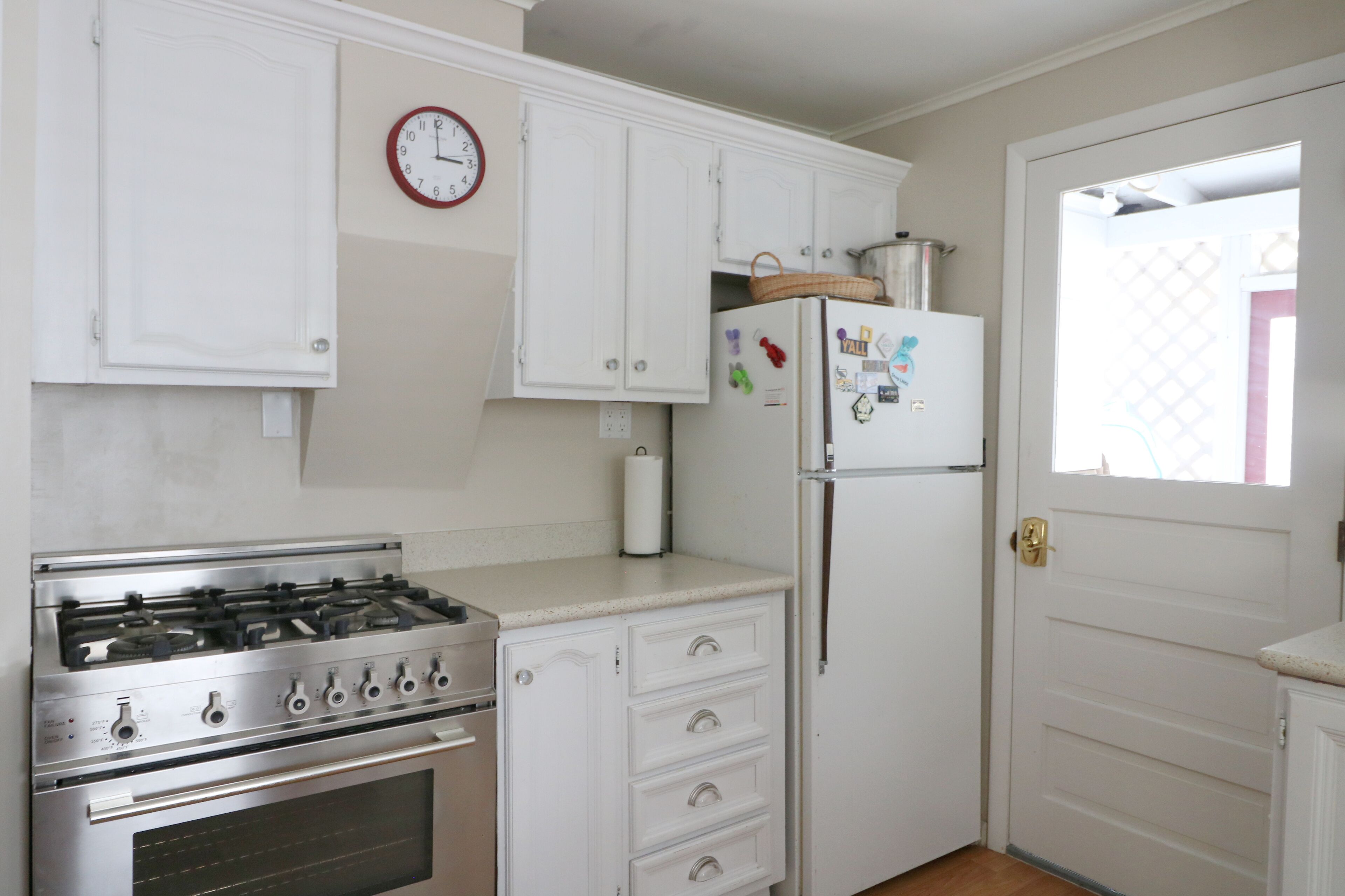 Une cuisine avec des meubles blancs, une cuisinière en acier inoxydable, un réfrigérateur orné d’aimants et une horloge au mur.