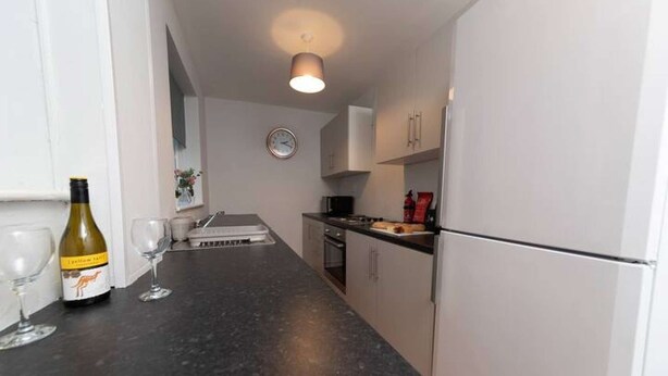 A small kitchen with a refrigerator, a microwave, and a sink. There is a wine bottle and glasses on the counter. - Hampton House Spacious Accommodation In Stockton (Stockton-on-tees)