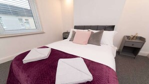 A neatly made bed with a purple blanket and white pillows, two bedside tables with lamps, and a window with blinds. - Hampton House Spacious Accommodation In Stockton (Stockton-on-tees)