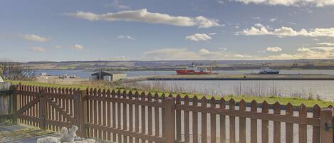 A wooden fence, a birdbath, and a body of water with boats in the distance.