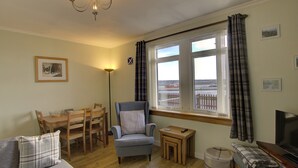Un salon chaleureux comprenant un coin repas, un fauteuil confortable, une petite table, un téléviseur et une vue sur une clôture par la fenêtre.