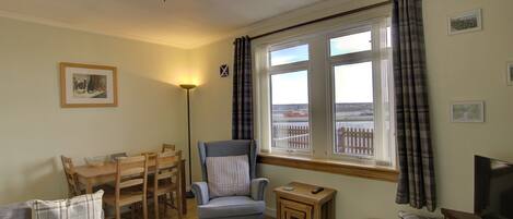 A cozy living room with a dining area, a comfortable armchair, a small table, a television, and a view of a fence through the window.