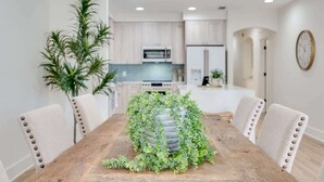 Un coin repas avec une table en bois entourée de chaises, un arrangement floral décoratif en guise de centre de table, et une vue sur une cuisine en arrière-plan.