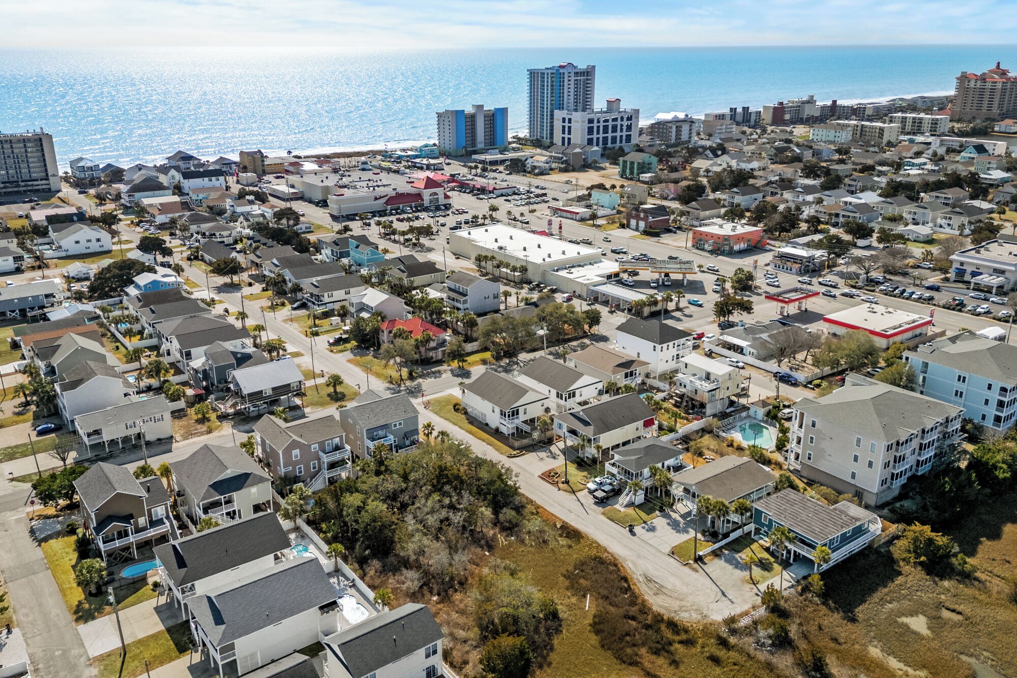 An aerial view of a coastal residential area with houses, a gas station, and a beach in the background.