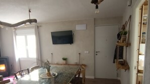 Ein Essbereich mit einem Glastisch, Holzstühlen, einem an der Wand befestigten Flachbildfernseher und einer Deckenleuchte.
