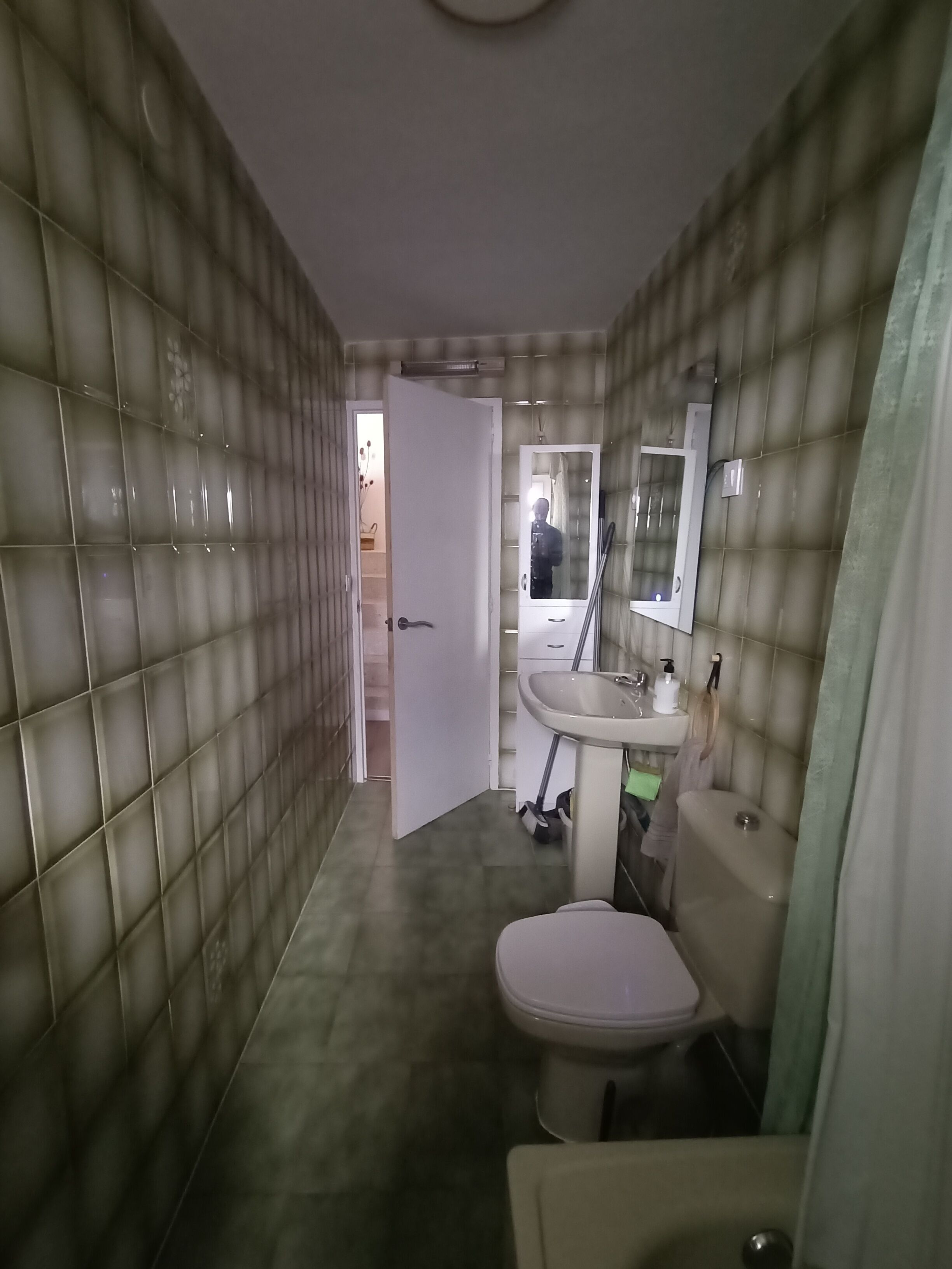 A bathroom with a toilet, sink, and mirror. The walls are tiled, and there is a door leading to another room.