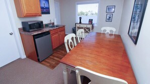 A compact kitchen with a microwave, sink, and wooden cabinets. There is a dining table with chairs and a window with blinds. - Egret Escape 2 Queen Deluxe Fin N Feather (Nags Head)